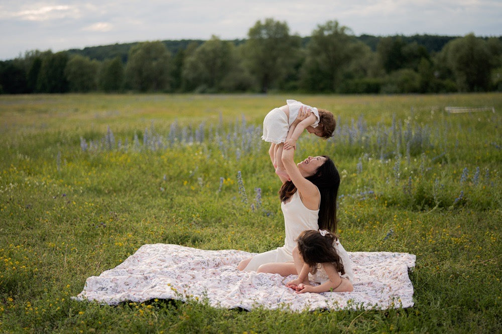 Moeder en twee jonge kinderen genieten van quality time op een grote, zachte picknickmat met bloemenmotief in een zonnig veld omringd door bloemen en bomen.