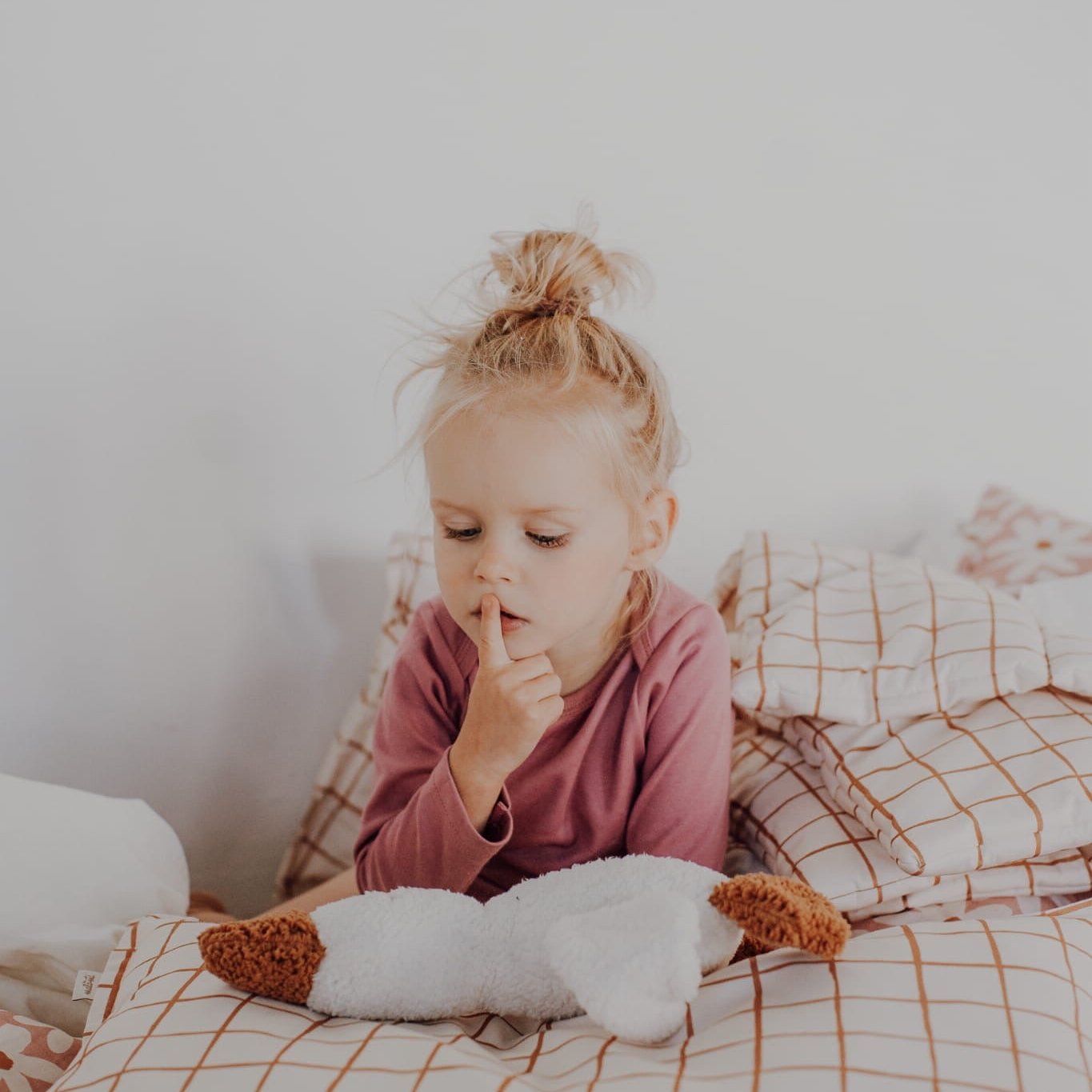 A child sitting on camel check bedding, holding a white plush goose toy, surrounded by cozy pillows