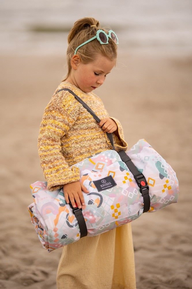 Klein meisje met zonnebril draagt een opgerolde, kleurrijke Makaszka picknickmat op het strand, klaar voor een dag vol plezier.