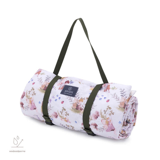 Opgerolde zachte picknickmat met sprookjesachtig bloemenpatroon, vastgezet met groene draagbanden en voorzien van een Makaszka-label, klaar om als schoudertas gedragen te worden.
