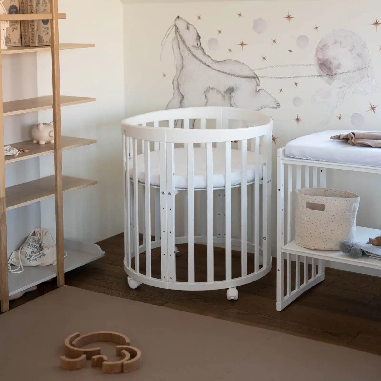 Klein wit rond babybedje in een minimalistisch ingerichte kamer met houten accenten. Het bedje staat naast een bijpassende commode met een zachte aankleedkussen.