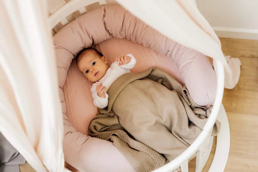 Baby ligt comfortabel in een rond houten babybedje met een zachte, roze bedomrander en een beige deken. Het bedje heeft een elegante baldakijn voor een knusse sfeer.