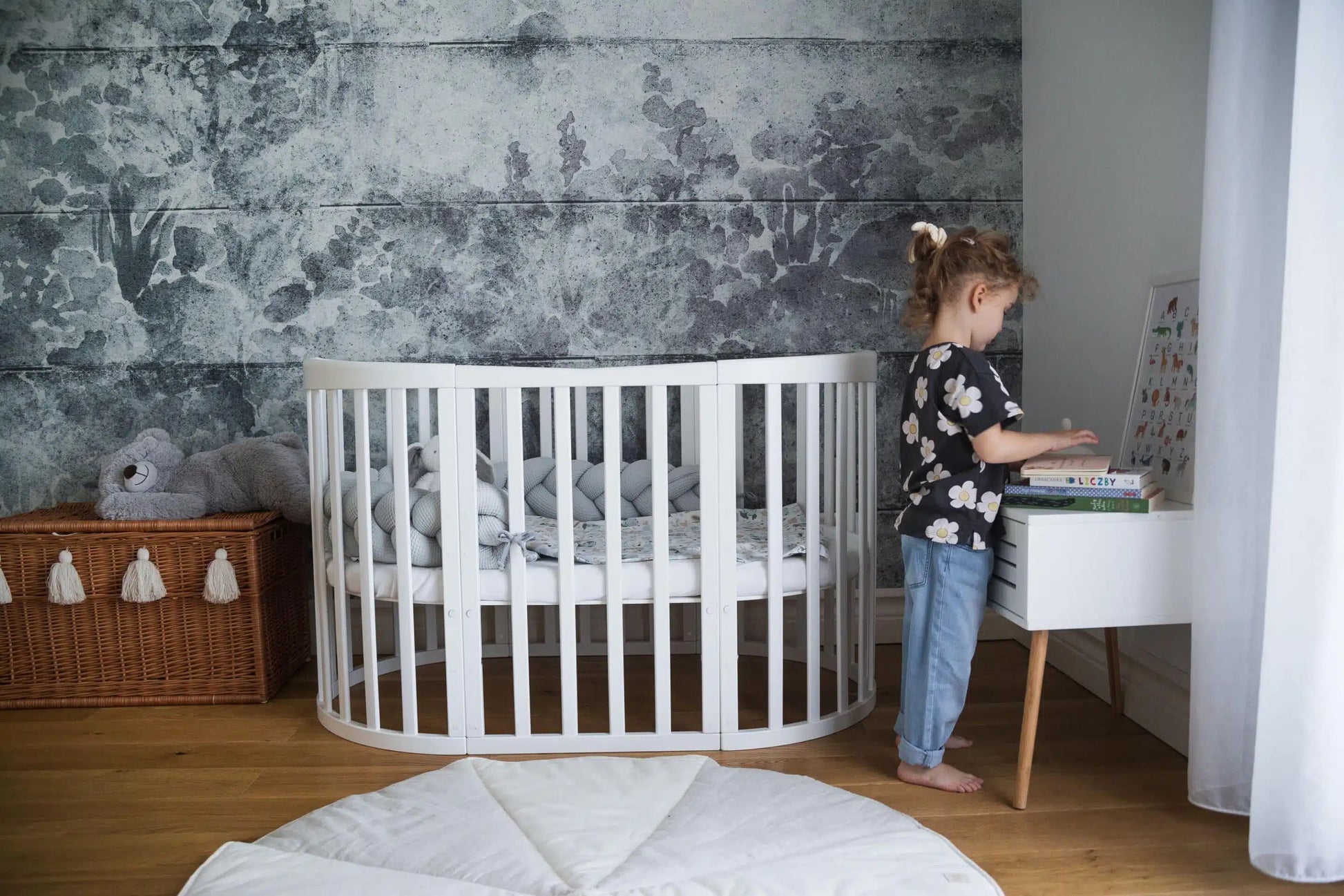 Wit houten babybed in een moderne kinderkamer met een grijze muur en een jong kind dat naast het bed boeken leest. Het bed is omgebouwd tot een groter babybed met een stijlvolle inrichting.