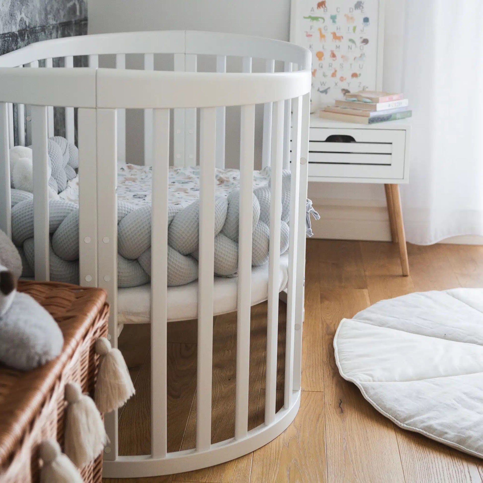 Wit houten babybedje in een stijlvolle Scandinavische kinderkamer, met een zachte gevlochten bedomrander en bijpassende decoraties. Het bedje is in een grotere stand gezet voor een peuter.