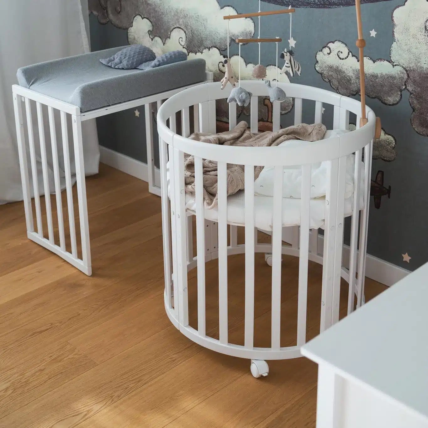 Rond wit houten wiegje met rubberen wielen, geplaatst in een babykamer met een sterrenhemelthema. Een mobiel met zachte figuren hangt boven het bed.