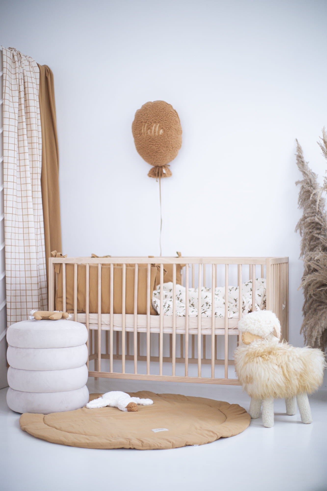 Een prachtige babykamer met natuurlijke tinten en zachte texturen. De ruimte bevat een houten ledikant, een ronde speelmat in warme camel kleur, een pluche schaapstoel, en bijpassende decoratieve elementen zoals een ballonwanddecoratie en kussens. Perfect voor een gezellige en stijlvolle kinderkamer.