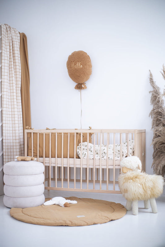 Een prachtige babykamer met natuurlijke tinten en zachte texturen. De ruimte bevat een houten ledikant, een ronde speelmat in warme camel kleur, een pluche schaapstoel, en bijpassende decoratieve elementen zoals een ballonwanddecoratie en kussens. Perfect voor een gezellige en stijlvolle kinderkamer.