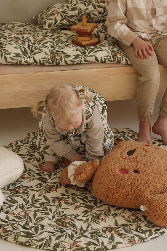 Een schattige baby speelt op een zachte Woodland speelmat met een prachtig blad- en dierenpatroon, naast een houten bed met bijpassend beddengoed. Een gezellige en stijlvolle kinderkamersetting die comfort en natuur combineert.